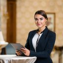 Photo of business woman in expensive hotel. Young business woman wearing suit, standing in nice hotel room, using tablet computer and looking at camera